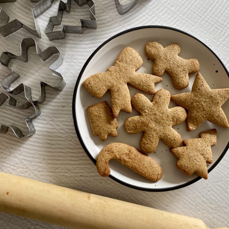 Christmas themed gingerbread cookies on a white plate with cookie cutters on the top left hand side and rolling pin at the bottom.